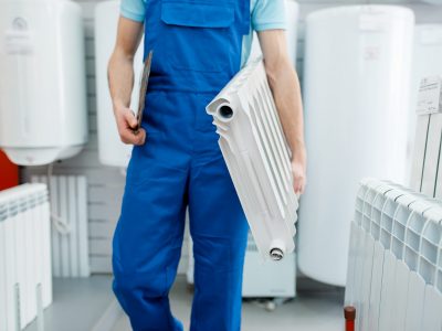 Plumber in uniform holds water heating radiator in plumbering store. Man buying sanitary engineering in shop